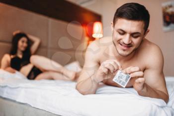 Love couple lying in bed, smiling man holds a condom in his hand. Intimate life, sex lovers in bedroom 