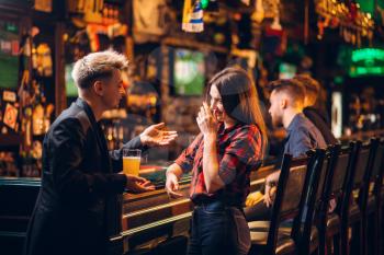 Young man holds glass of beer in hand and talks with woman at the bar counter in a sport pub, happy leisure of football fans