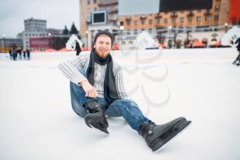 Young smiling man in skates sitting on ice, skating rink. Winter ice-skating on open air, active leisure