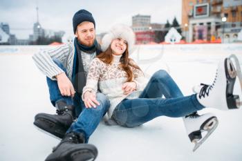Young love couple in skates sitting on ice, skating rink. Winter ice-skating on open air, active leisure