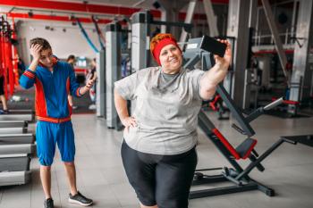 Fat woman makes selfie with instructor in gym, humor. Calories burning, obese female person in sport club, fat-burning