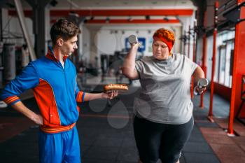 Trainer with hotdog in hand forces fat woman to exercise, motivation, hard workout in gym. Calories burning, obese female person in fitness club, fat-burning, sport against unhealthy food