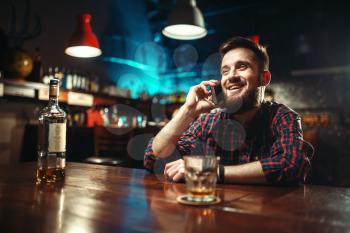 Smiling man sitting at the bar counter and talking by phone, relaxation with alcohol. Happy guy having fun, leisure in pub