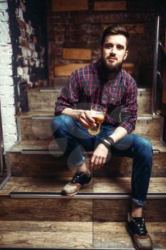 One bearded man sitting on the stairs in bar and holds glass of beer in hands. Male person leisure in pub