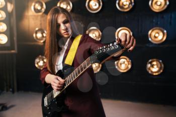 Female musician in suit playing on electric guitar, stage with lights on background, retro style. Live music performer, rock guitarist