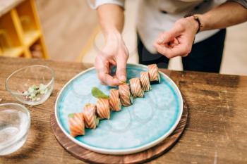 Male person cooking sushi rolls with salmon on wooden table, japanese food preparation process.