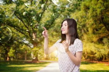 Cute woman catches soap bubble, summer park on background. Female person blowing colorful balloons