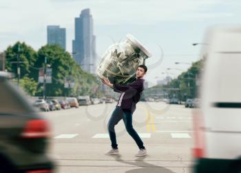 Money saving concept, young man trying to carry a big glass jar full of dollars. Cash economy addiction, home bank