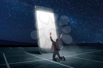 Phone addicted people concept. Man praying on his knees against large smartphone with glowing screen. Scaling effect