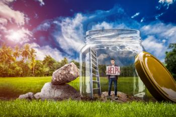 Fear of loneliness concept, young helpless man with help sign standing in a big glass jar on the green meadow. Depression of alone people