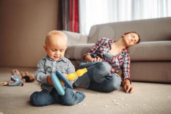 Little kid playing on the floor, mother sleeps, motherhood problems. Stressed mom and son together at home, parenthood. 