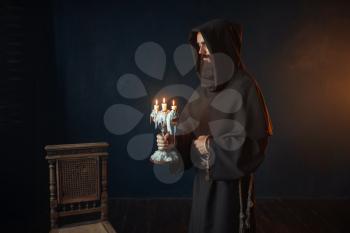 Medieval monk in robe holds a candlestick in hands, black background, secret ritual. Mysterious friar with candles. Mystery and spirituality