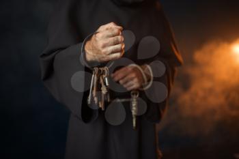 Medieval monk in robe holds a bunch of keys in hands, black background, secret ritual. Mysterious friar in dark cape. Mystery and spirituality