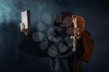 Medieval monk stands in fire with book in hands, black background, secret ritual. Mysterious friar in dark cape. Mystery and spirituality