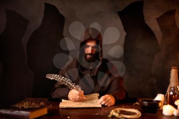 Medieval monk in robe makes notes in the secret scripture with a goose feather, black background, secret ritual. Mysterious friar in dark cape. Mystery and spirituality