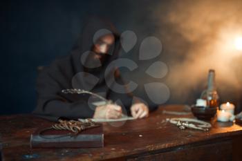 Medieval monk in robe and hood writes with a goose feather, black background, secret ritual. Mysterious friar in dark cape. Mystery and spirituality
