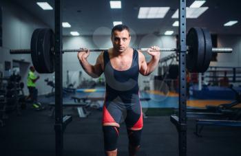 Muscular athlete in sportswear poses at the stand with barbell in gym. Weightlifting workout, lifting