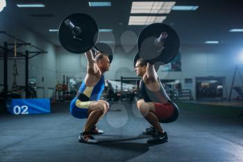 Two male weightlifters doing squats with barbells, gym interior on background. Weightlifting workout in sport or fitness club, bodybuilding