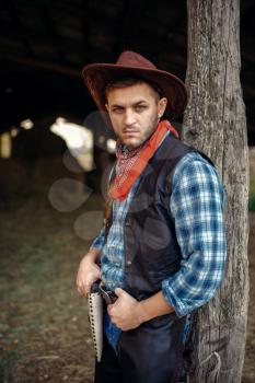 Brutal cowboy in jeans and leather jacket, texas ranch on background, western. Vintage male person with revolver, wild west lifestyle