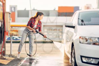 Cute woman cleans car wheels with high pressure water gun. Young lady on self-service automobile wash. Outdoor vehicle washing at summer day 
