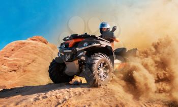 Quad bike in dust clouds, sand quarry on background. Male driver in helmet on atv in desert dunes