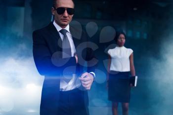 Serious bodyguard in suit, sunglasses and earpiece, female client on background. Security guard is a risky profession, professional guarding of business persons