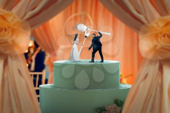 Wedding cake, bride and groom figurines hold huge cutlery in their hands on the top. Bridal pie for newlyweds with little figures, traditional celebration ceremony symbol