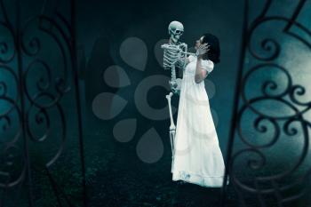 Female victim with human skeleton at the gates of the old cemetery at the night. Photo in horror style, exorcism