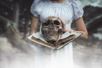 Female victim in white dress holds book and human skull in hand, forest on background. Photo in horror style, exorcism