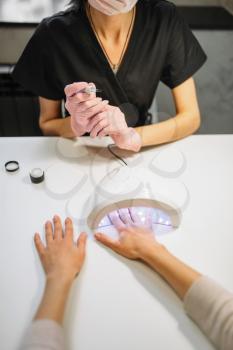 Female client dries gel varnish in the nail dryer, beautician in pink gloves on background, beauty salon. Manicurist doing hands care cosmetic procedure