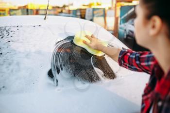 Young woman with sponge scrubbing vehicle glass with foam, car wash. Lady on self-service automobile washing. Outdoor carwash