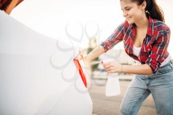 Cute woman cleans rear lights of the car with sponge and spray, carwash. Lady on self-service automobile washing. Outdoor vehicle cleaning at summer day 