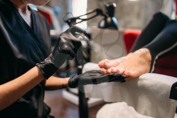 Pedicure master in black gloves sprays foot nails of female client, beauty salon. Professional  fingernail care