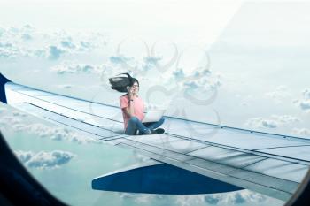 Communication addiction, woman with laptop and phone sitting on plane wing, clouds on background