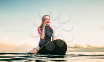 Female sportman windsurfer in wetsuit sitting on the board and talks by phone