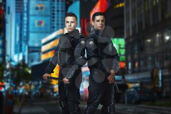 Two cops with a gun and a baton on the guard of the law, concept. Special force troops in black body armor, front view, night cityscape on background