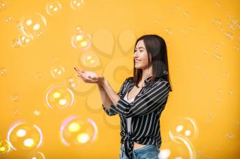Pretty woman catches soap bubble, yellow background. Female person blowing colorful balloons