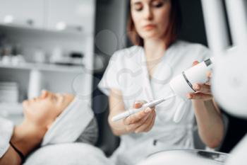 Beautician prepares laser for rejuvenation procedure, female patient on background, cosmetology clinic. Facial skincare in spa salon