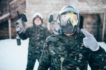 Paintball player points finger at splattered mask, team after winter battle. Extreme sport game, soldiers in special uniform, paintballing