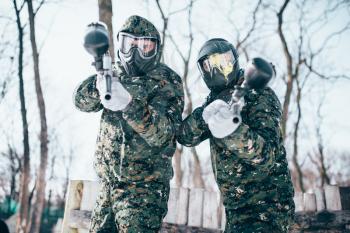 Two paintball players in splattered masks, team poses after winter battle. Extreme sport game, soldiers in special uniform, paintballing