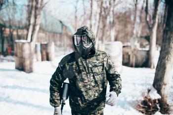 Male paintball player in protection mask and uniform holds marker gun in hands, soldier before winter forest battle. Extreme sport, military game equipment