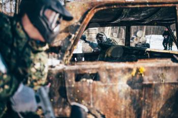 Paintball battle, players fight around burned car in winter forest, paintballing. Extreme sport, military game
