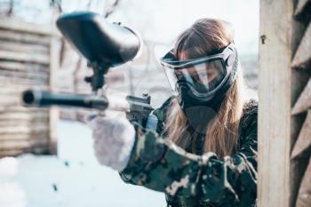 Female paintball player with marker gun in hands, winter forest battle. Extreme sport game, woman fights in protection mask and uniform