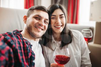 Love couple sitting on the floor against couch, watches movie and drinks red wine from big glasses, window and living room interior on background. Romantic evening