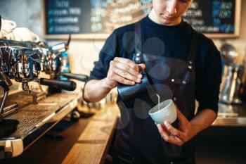 Barista in apron pours cream into the cup of coffee, cafe counter on background. Professional cappuccino preparation by bartender