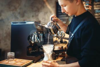 Male barista pours hot water into the glass with coffee, cafe counter and espresso machine on background. Barman works in cafeteria, bartender occupation
