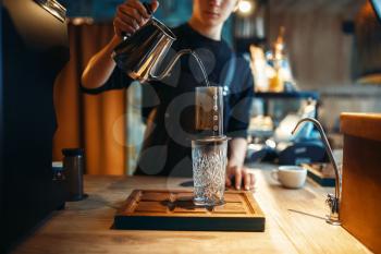 Male barista pours hot water from coffee pot into the glass, cafe counter and espresso machine on background. Barman works in cafeteria, bartender occupation
