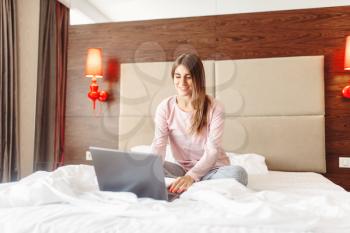 Smiling woman sitting in bed and uses laptop, good morning, bedroom interoir on background