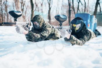 Two paintball players lies on the snow and shooting at the enemy in winter forest battle. Extreme sport game
