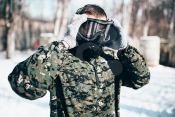 Male paintball player puts on protection mask before winter forest battle. Extreme sport, military game equipment